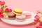 Plate of macaroons on pink background with flowers. Sweet pastry, baked products, sweets, dessert. Unhealthy diet