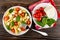 Plate with pasta, tomatoes cherry, feta and spinach, fork, plate with pieces of cheese, spinach and tomatoes on napkin on wooden