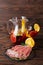 A plate of prosciutto and ham, a glass jug of beverage with apples and oranges on a wooden brown background.