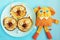 A plate with three pancakes in the shape of cat faces, decorated with slices of banana, grape and apple. orange toy dog.