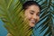 Playful girl peeks out from behind palm leaves, smiles.