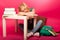 Playful schoolgirl with two hair tails and big eyeglasses sitting behind the small table full of books fall asleep while studying.