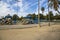 A playground with a blue and brown jungle gym and a baseball field with lush green palm trees and grass with blue sky and clouds