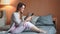 Pleasant barefoot female chatting using smartphone sitting on bed relaxing at cosiness home