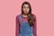 Pleasant looking brunette woman pouts lips, makes grimace at camera, wears purple jumper and denim overalls, wants to