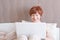 Pleasant senior woman with short red hair is sitting on the bed with a laptop, looking at the monitor and smiling