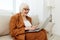 a pleasant, wise elderly woman is sitting looking at a laptop on the sofa in a relaxed pose in a bright room near the