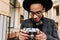 Pleased black man in headphones walking down the street with camera. Outdoor photo of curious male african photographer