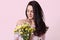 Pleased delighted dark haired young woman smells pleasant odour from flowers, dressed in stylish clothes, has healthy skin, models