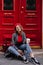 Pleased girl in glasses and black gumshoes drinks coffee, sitting near red door. Stunning young woman in beret posing on