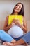 Pleased pregnant woman holding book while sitting on sofa