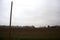 Ploughed field with a grove and buildings in the distance framed by a over head cable and a wooden pole on a cloudy day in the