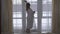 Plump girl in white long bathrobe walks to large floor-to-ceiling window with cup of coffee or tea. Lady straightening