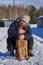 A plump middle-aged blonde woman hugs a mongrel sitting on white snow on a sunny winter day.
