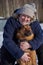 A plump middle-aged blonde woman hugs a mongrel sitting on white snow on a sunny winter day.