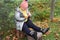 Plump smiling girl sits on stump outdoors in autumn day and cleans orange