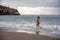 A plump woman in a bathing suit enters the water during the surf. Alone on the beach, Gray sky in the clouds, swimming