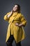 Plus size fashion model in yellow coat and black hat, fat woman on gray background, overweight female body