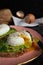 Poached egg cut with a knife on avocado puree on toast bread on a pink plate. Egg scarlop, bread and towel on background