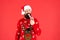 pondering bearded man in santa claus hat and sweater hold candy stick on red background, christmas