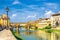 Ponte Vecchio stone bridge with colourful buildings houses over Arno River blue reflecting water and embankment promenade in histo