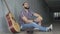 Poor Caucasian man sitting with guitar and hat in urban underground crossing and eating bun. Wide shot portrait of male