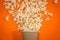 Popcorn spills out of the glass on an orange background, viewed from above. A splash of popcorn