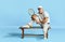 Popular game. Portrait of handsome senior man in stylish white outfit sitting on bench over blue background. Concept of