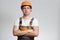 Poratrait of confident young construction worker in hard hat and overalls with arms folded on chest on grey background