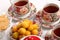 Porcelain teacups with hot tea and with floral pattern, teapot, biscuits and plate with wild apricots on a table