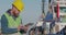 Port Worker With A Beard In A Yellow Helmet Stands With A Tablet PC In The Seaport Against The Background Of Ship. The Docker