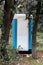 Portable blue and white ecological toilet behind tall trees
