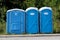 Portable blue and white ecological toilets in a row for men woman and disabled persons next to paved road with trees in background