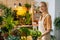 Portait of smiling young woman florist in apron spraying water on houseplants in pots by sprayer, looking at camera.