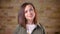 Portait of young attractive brown-headed woman watching smilingly into camera on bricken wall background.