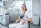 Portraait of sincerely smiling young dentist woman dressed white medical scrubs uniform sitting in modern dental clinic next to