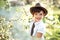 Portrait of 7 years old cute stylish short haired smiling girl in white shirt and brown hat looking away in blooming garden in