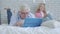 Portrait of absorbed Caucasian mid-adult man using laptop as blurred woman with smartphone sitting on bed at the