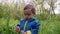 Portrait of adorable baby boy playing with dandelion on green forest background. Cute kid in blue raincoat explores