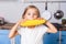 Portrait of adorable cute child girl biting big vegetable corn, standing in modern kitchen, healthy nutrition without GMOs. indoor