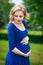 Portrait of adorable pensive young pregnant woman in blue dress with long blond curly hair holding her belly in summer park