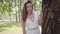Portrait adorable young girl with long brunette hair wearing a long white summer fashion dress standing next to a tree