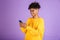 Portrait of african american man smiling and holding smartphone, wearing bluetooth earphone, isolated over violet background