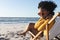 Portrait of african american woman in sunglasses sitting in deckchair smiling on sunny beach by sea