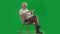 Portrait of aged bearded man on chroma key green screen. Full shot senior man sitting on chair surfing web on tablet.