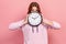 Portrait of amazed curly haired teenage girl in hoodie peeping behind big wall clock, time management and deadline