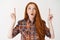 Portrait of amazed redhead girl pointing fingers up, looking at logo with interest, standing over white background