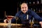 Portrait of angry and stressed barman with bowtie behind the bar with alcoholic drinks around. Stressful lifestyle of