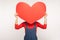 Portrait of anonymous girl in denim overalls hiding face behind big red paper heart, holding huge greeting card