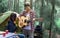 Portrait of Asian man wearing check shirt and hat playing guitar and singing for his beautiful girlfriend while camping in